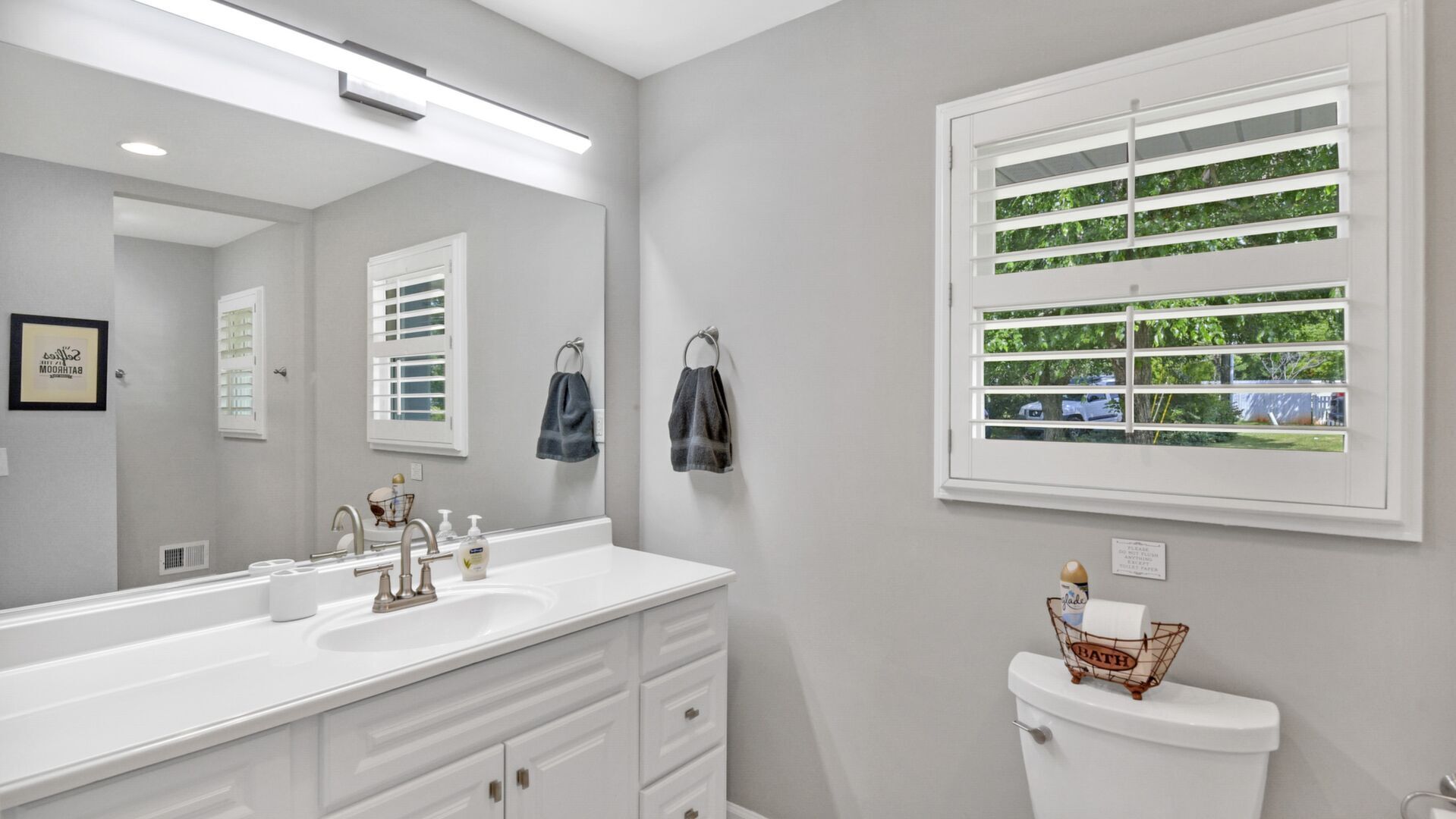 A bathroom with a white vanity, a large mirror, a window with blinds, and a toilet with a basket labeled "BATH" on top.