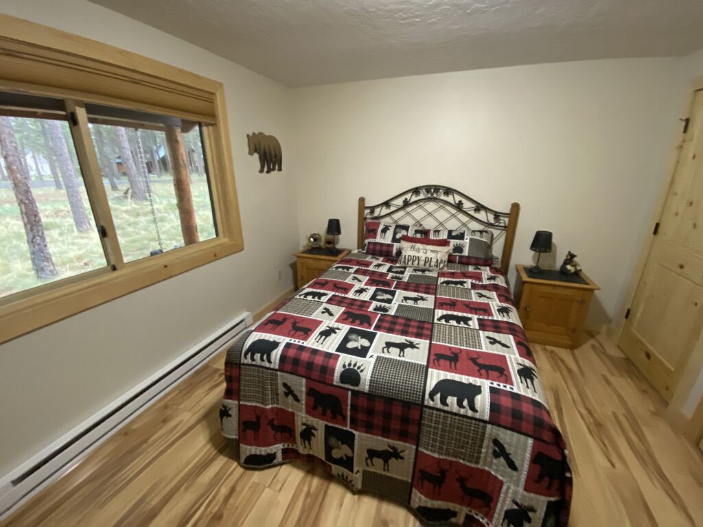 Een slaapkamer met een houten bed, een raam met uitzicht op bomen, een nachtkastje met een lamp en een deur.