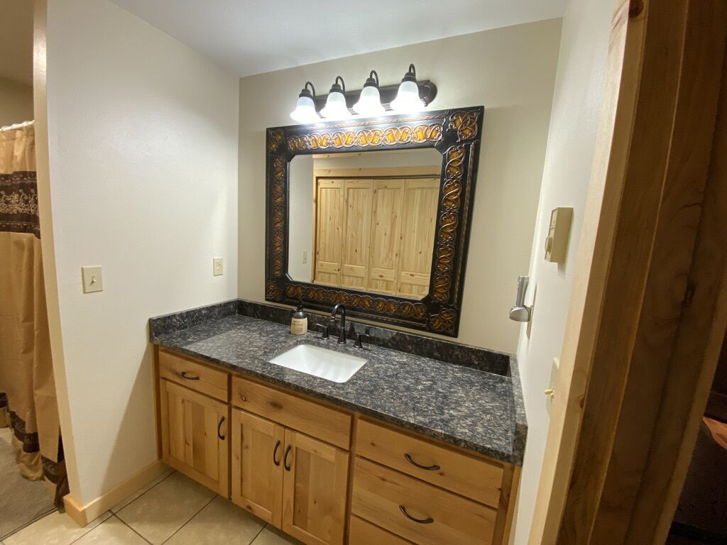 A bathroom with a granite countertop sink, wooden cabinets, a large mirror with decorative frame, and wall-mounted lights.