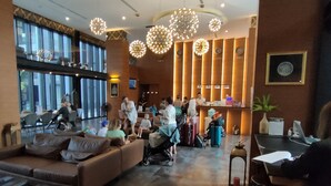 Un hall d’entrée spacieux avec un plafond haut, de grandes fenêtres et un coin salon avec des canapés. On y trouve plusieurs luminaires et un comptoir d’accueil.
