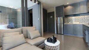 Un salon moderne avec un canapé et une table basse, une cuisine équipée d’un réfrigérateur et d’un plan de travail recouvert de carreaux, et une porte vitrée donnant sur une autre pièce.