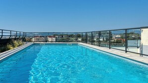A rooftop swimming pool with clear blue water, surrounded by glass railings and a view of buildings in the background.