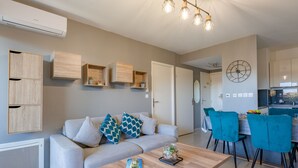 A modern living room with a sofa, coffee table, and teal chairs. There is a kitchen area with a dining table and chairs.