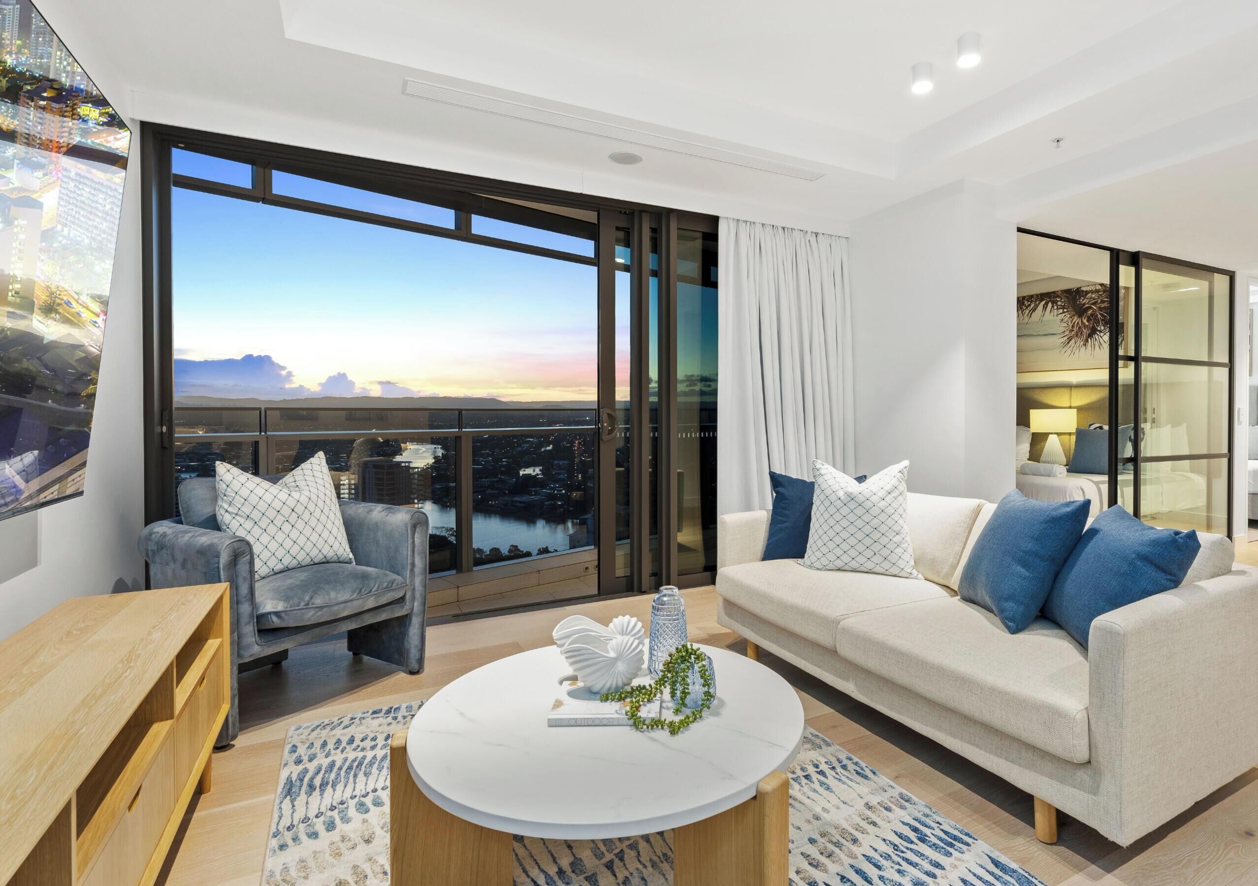 A modern living room with a large window, a sofa, a coffee table, and a view of a cityscape.