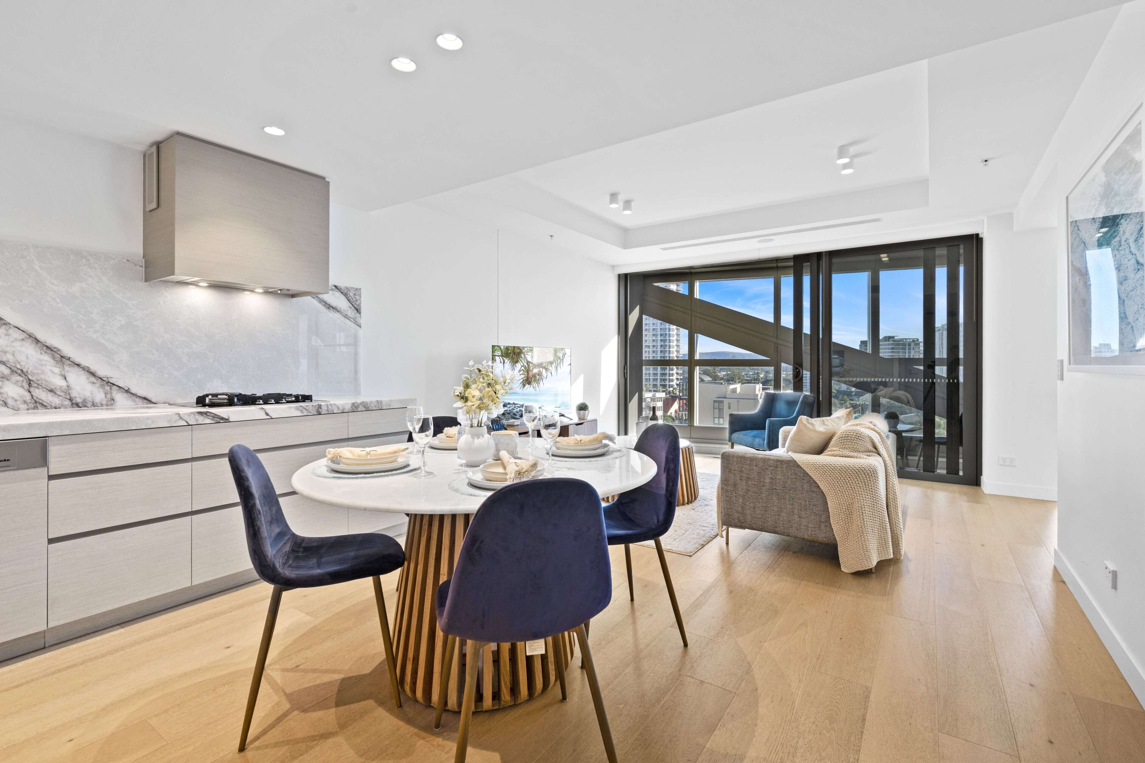 A modern kitchen with a marble countertop, a built-in stove, and a dining area with a round table and blue chairs.