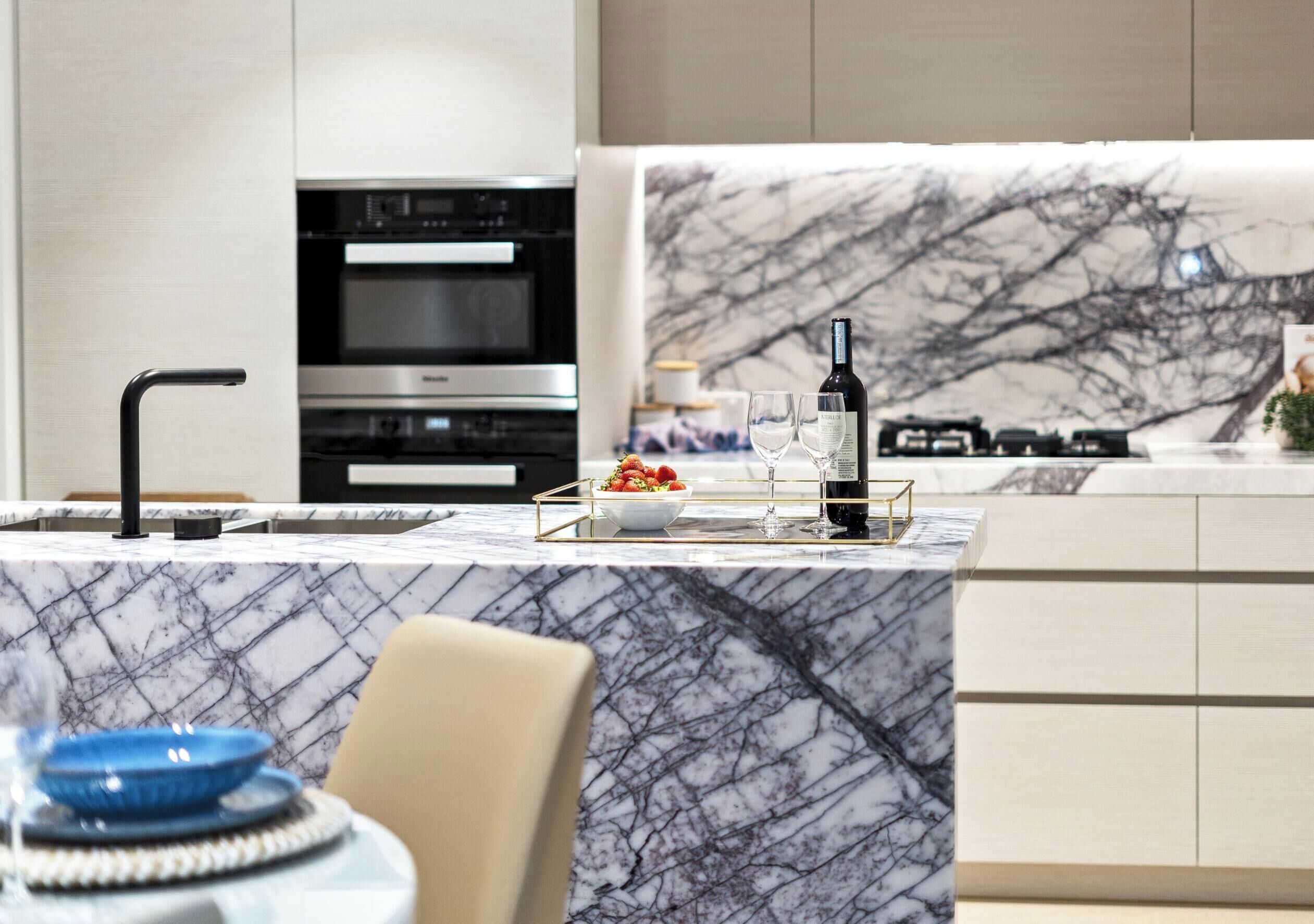 A modern kitchen with a marble countertop, stainless steel appliances, and a dining area with a chair and a blue bowl.