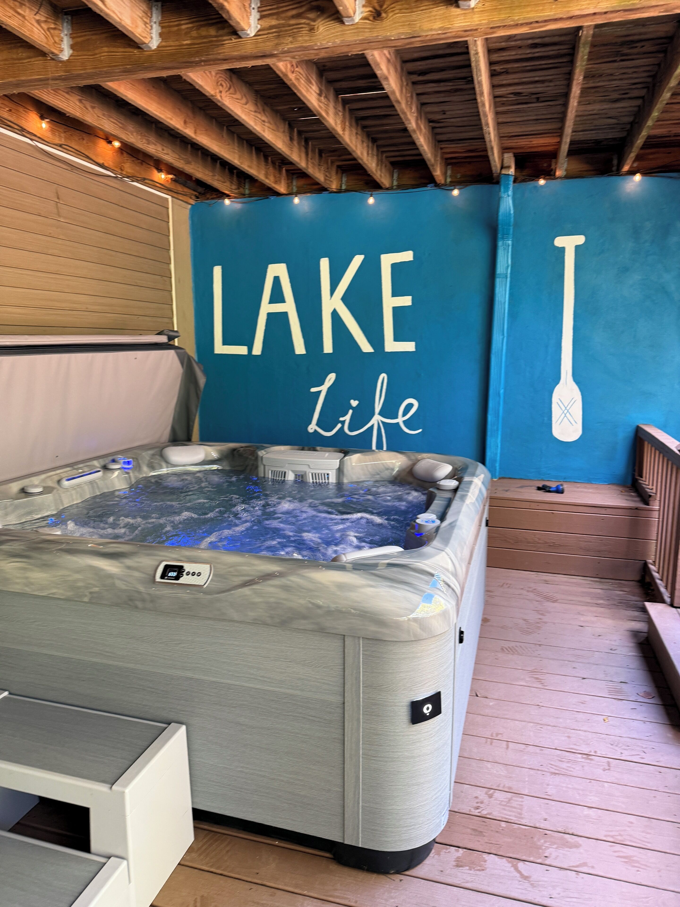 Ein Whirlpool mit digitaler Temperaturanzeige, eine Holzterrasse und eine Wand mit der Aufschrift 'LAKE Life' sowie ein Paddel.