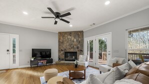 A spacious living room with a stone fireplace, a ceiling fan, and a large TV. - Lake Lanier Retreat + Deep Water Dock + Hot Tub (Gainesville)