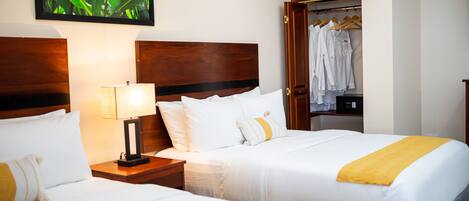 A hotel room with two beds, a wooden headboard, a nightstand with a lamp, and a wardrobe with hanging clothes.