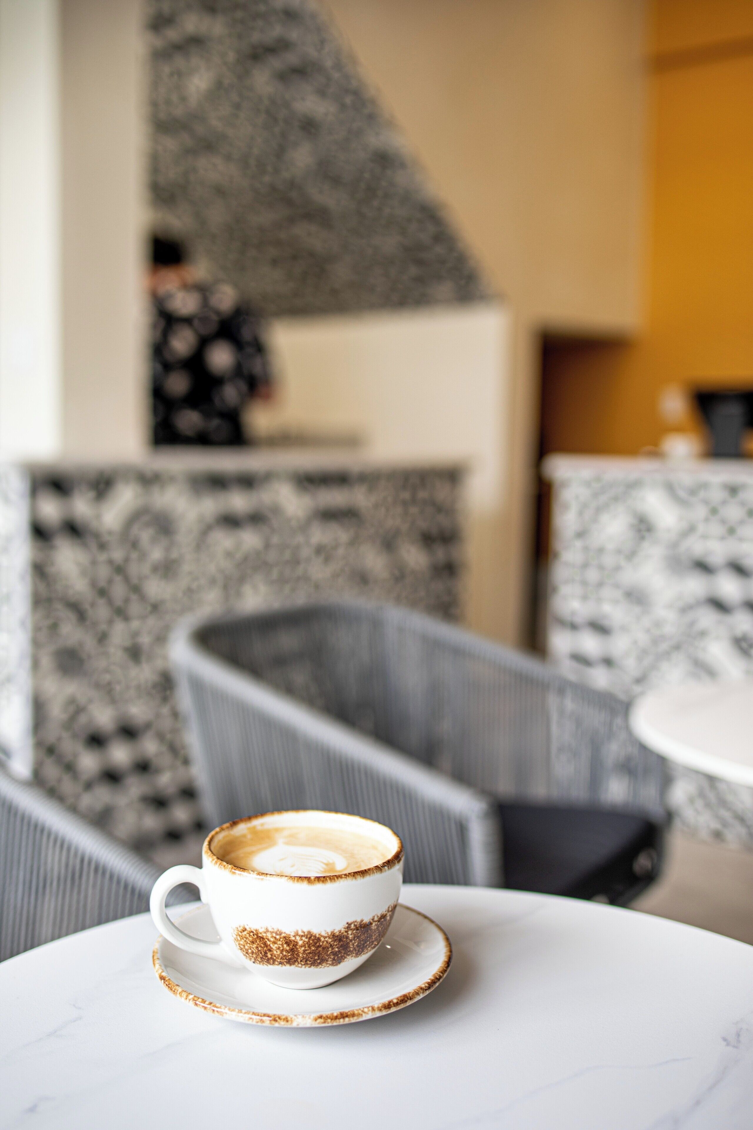 A cup of coffee on a white table with a marble pattern. In the background, there is a person sitting on a chair, and a staircase with a patterned wall.