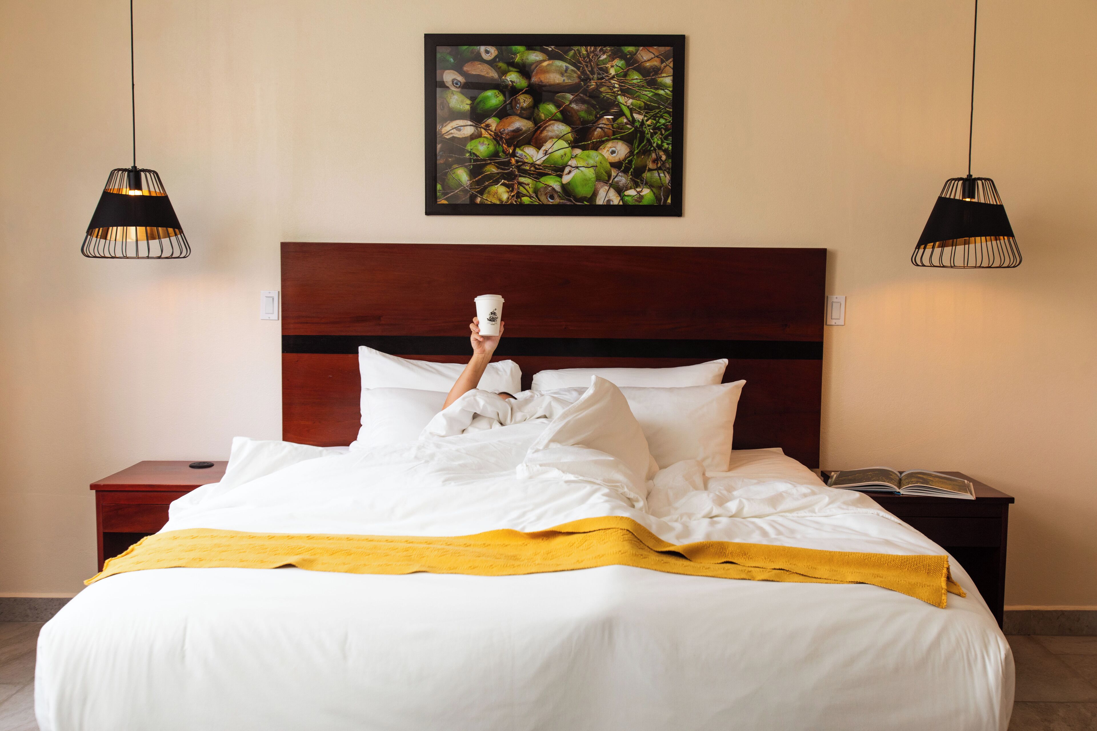 A neatly made bed with white linens and a yellow throw blanket, a wooden headboard, and two black pendant lights.