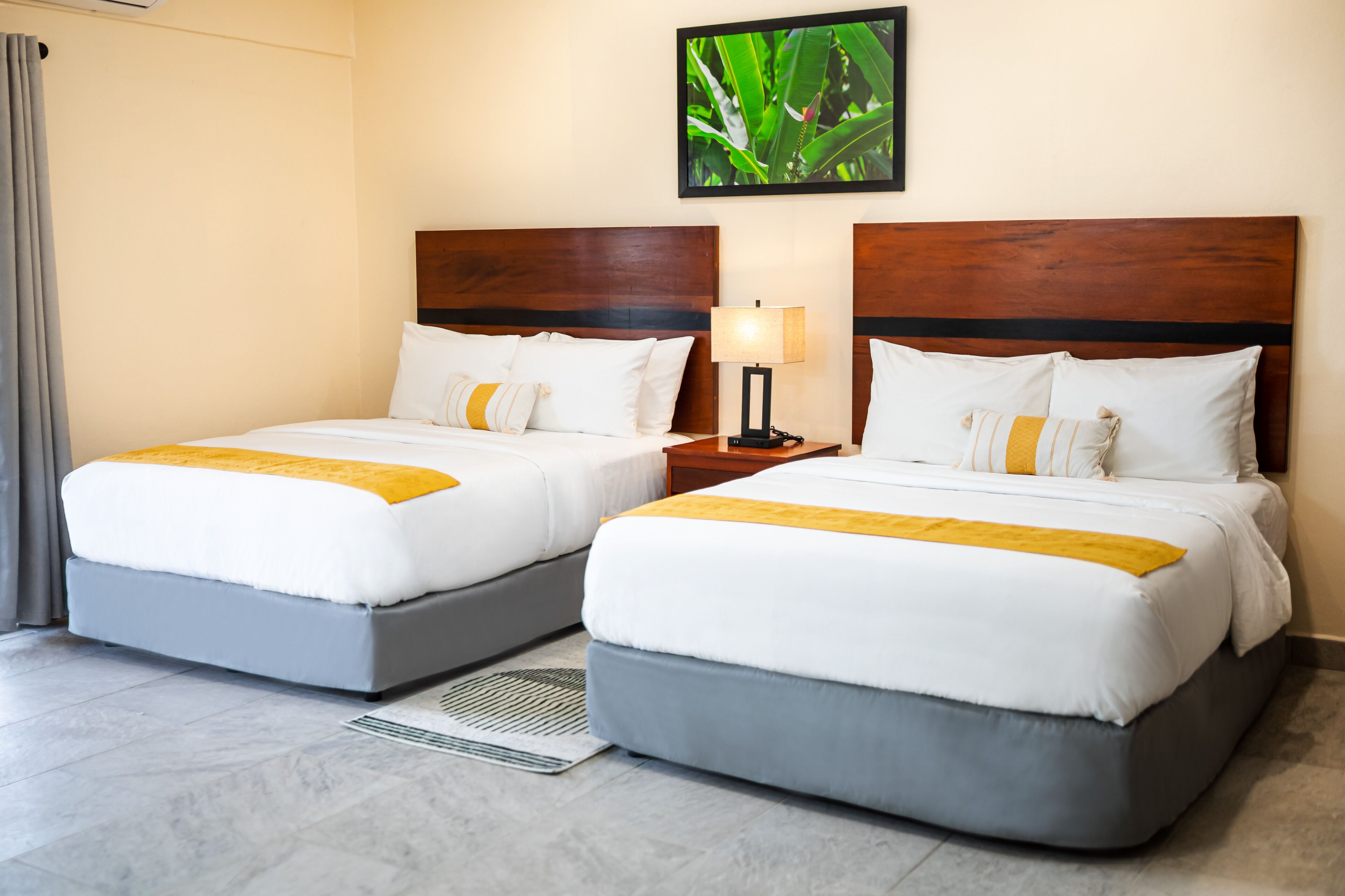 Habitación de hotel con dos camas, cada una con cabecero de madera, ropa de cama blanca y un toque amarillo. Hay una mesita de noche con una lámpara entre las camas, y se ve una planta en la pared.