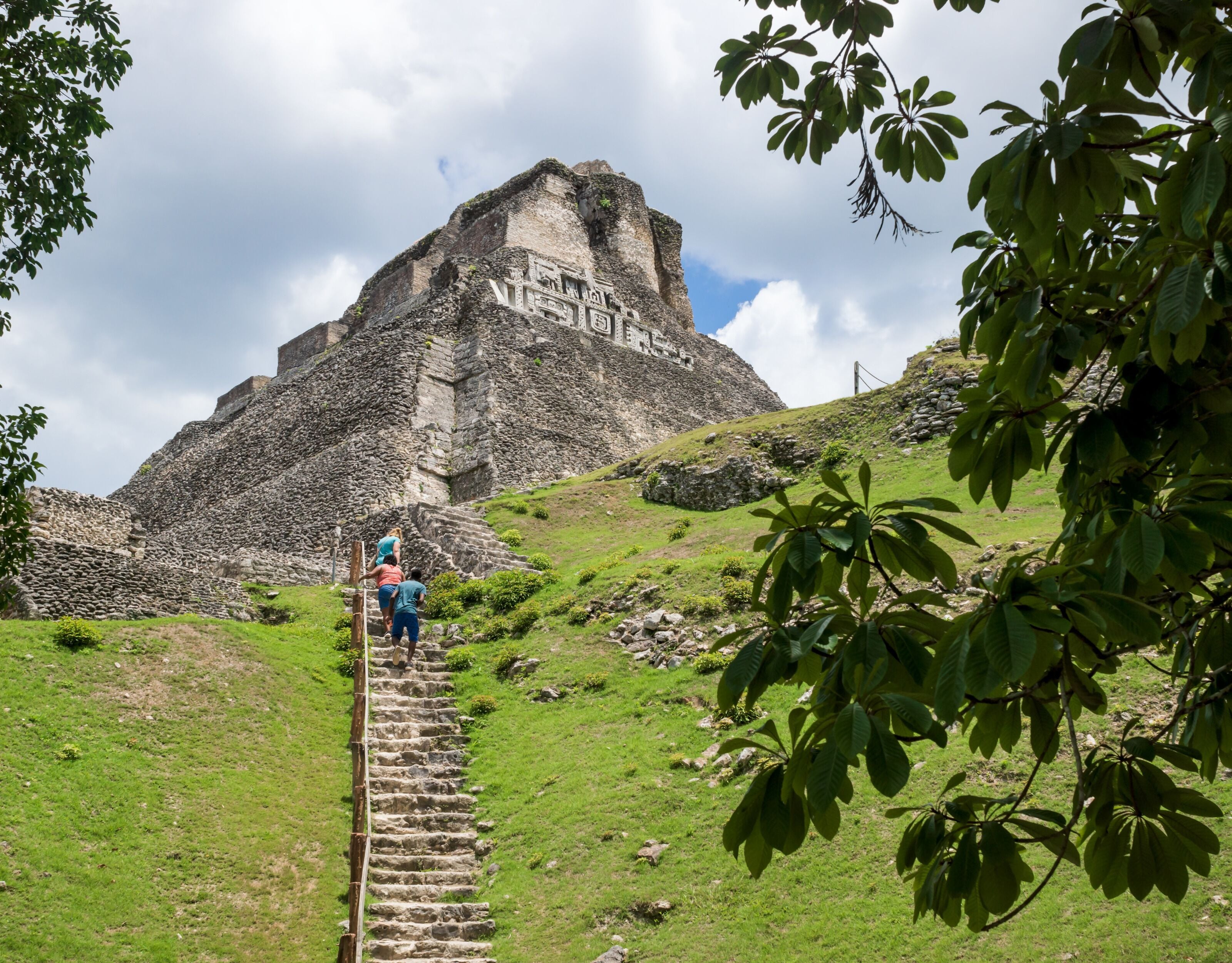A stone staircase leading up to a large pyramid-like structure with carvings, surrounded by greenery and a cloudy sky.