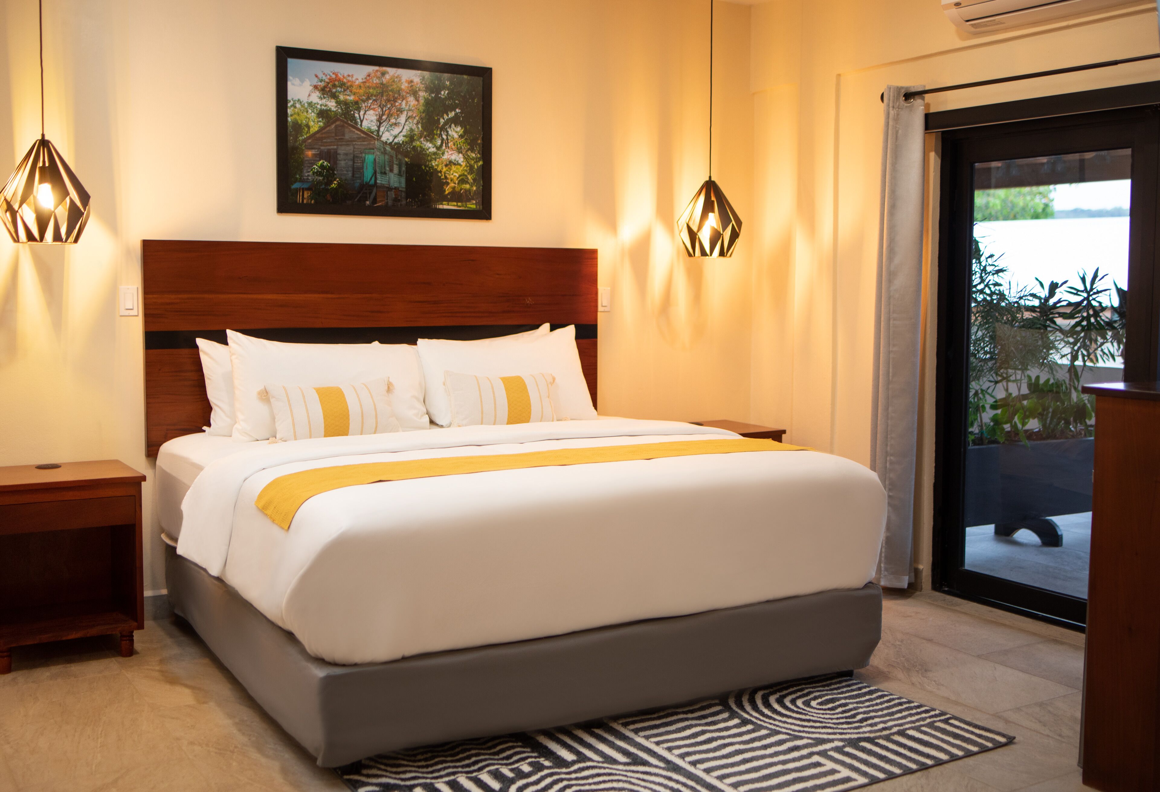 A neatly made bed with white and yellow bedding, two bedside tables, a framed picture on the wall, and a view of the outdoors through a glass door.