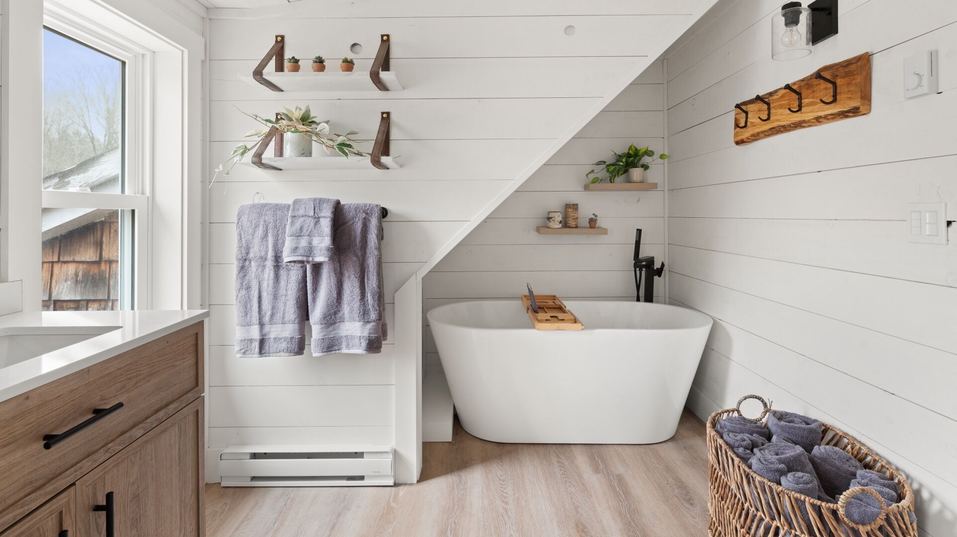 Un baño moderno con una bañera independiente, estantes de madera con plantas y una cesta de mimbre con toallas.