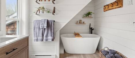 Un baño moderno con una bañera independiente, estantes de madera con plantas y una cesta de mimbre con toallas.