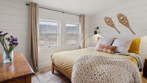 Et soveværelse med seng, et natbord, en lampe, to vinduer og udsigt til det fri.