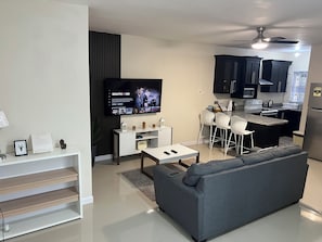 Una sala de estar moderna con un sofá gris, un televisor, un comedor con sillas blancas y una cocina con armarios oscuros.