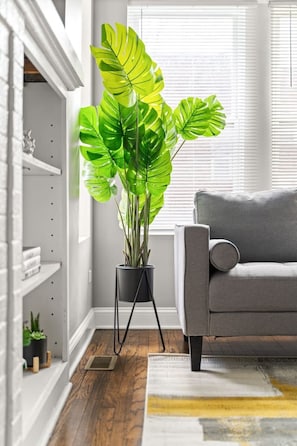 Una sala de estar con un sofá gris, una planta grande de hojas verdes en una maceta negra y una chimenea blanca.