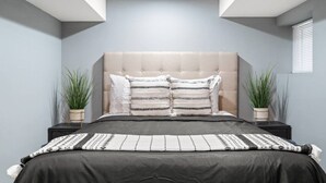Een moderne slaapkamer met een gestoffeerd hoofdeinde, twee nachtkastjes met potplanten en een groot bed met gestreept beddengoed.