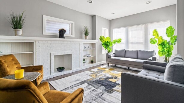Een moderne woonkamer met een open haard, een bank en fauteuils, met een wit bakstenen haard, een grijze bank en gele fauteuils.