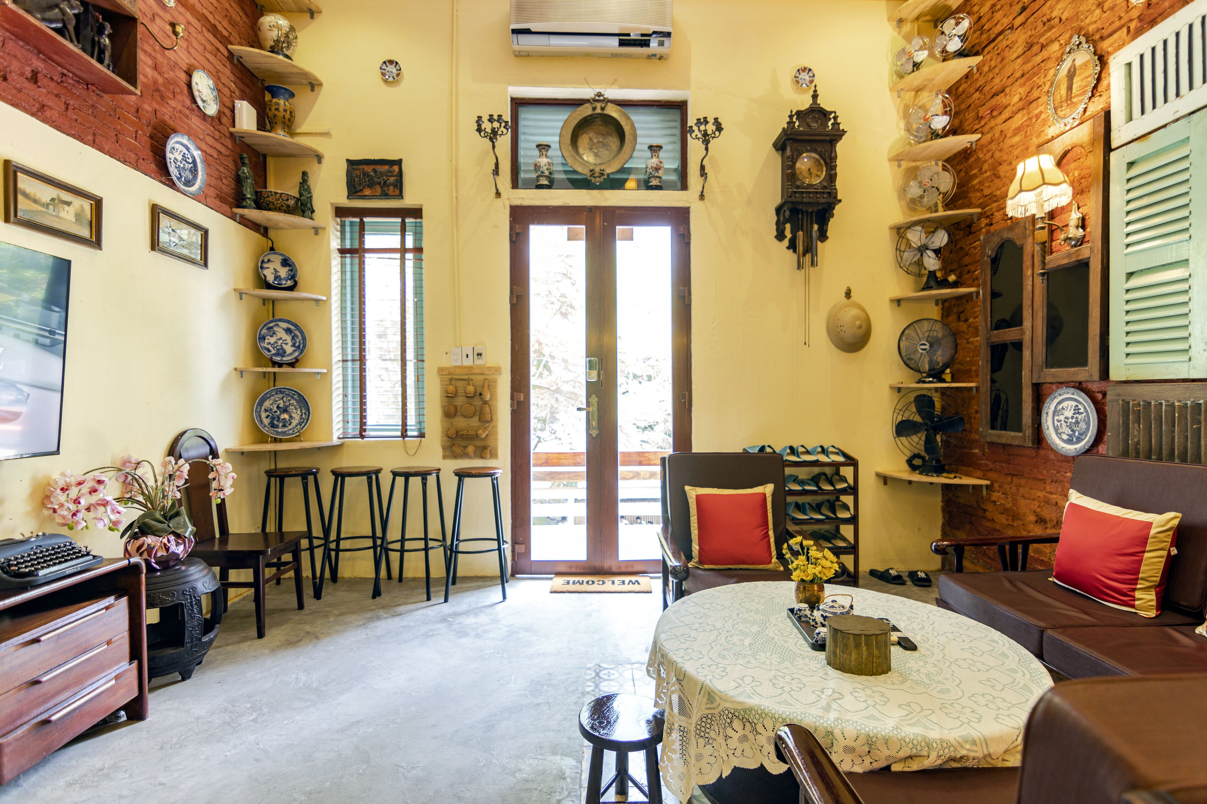 A cozy room with a round table, bar stools, a sofa, and a chair. There are shelves with decorative plates and a clock on the wall.
