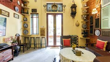 A cozy room with a round table, bar stools, a sofa, and a chair. There are shelves with decorative plates and a clock on the wall.