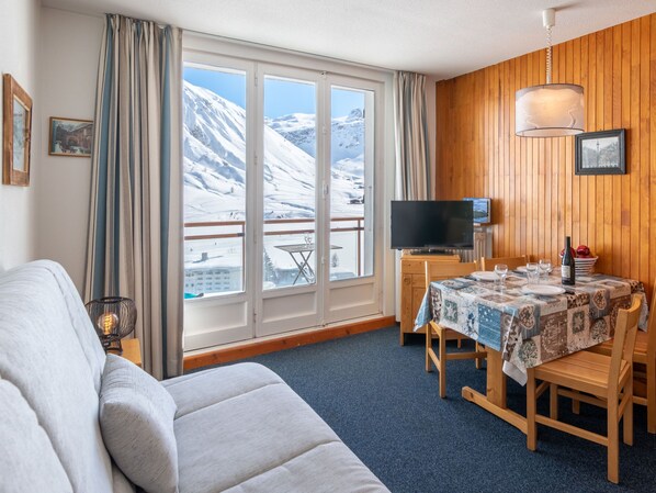 A cozy room with a wooden interior, a large window with a snowy mountain view, a dining table set with plates and glasses, and a flat-screen TV on a wooden stand.