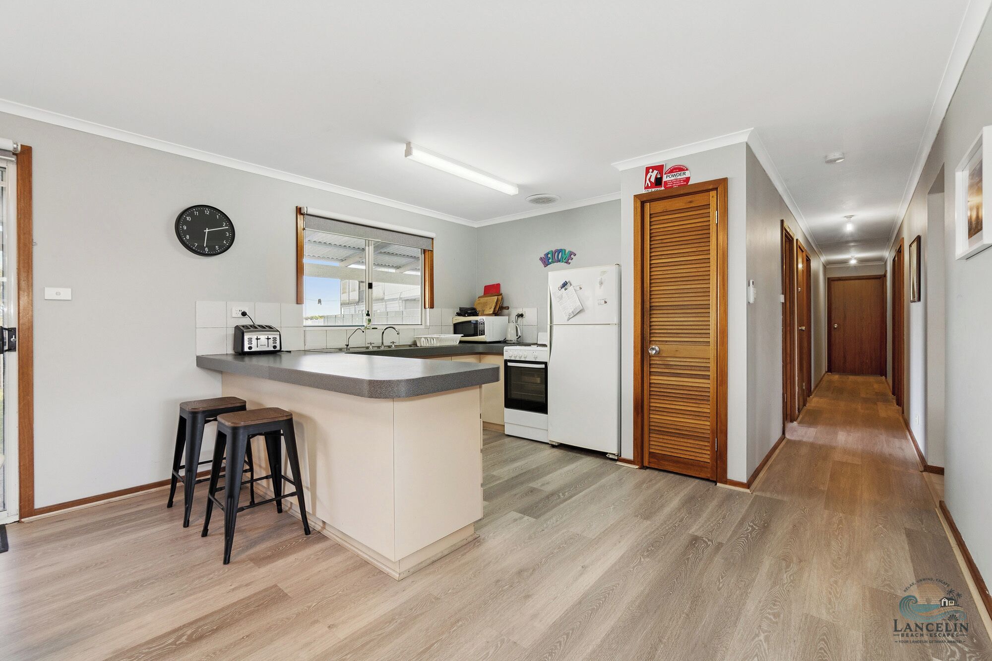 A kitchen with a central island, a microwave, a toaster, and a refrigerator. There is a clock on the wall and a wooden door with a 'Welcome' sign.