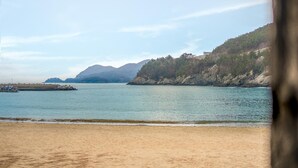 Ein Strand mit sandigem Ufer, ruhigem Wasser und einer bewaldeten Hügelkette im Hintergrund.