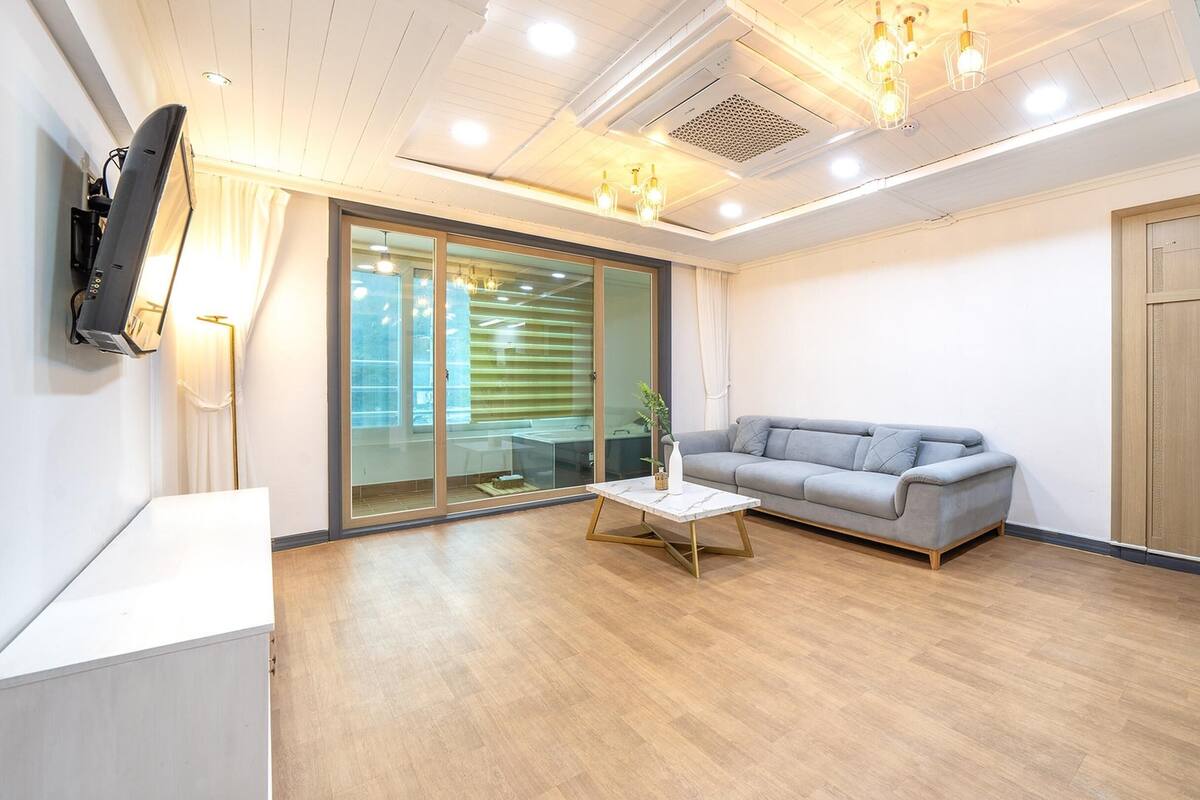 Een moderne woonkamer met een grijze bank, een klein wit tafeltje en een keuken met een koelkast.