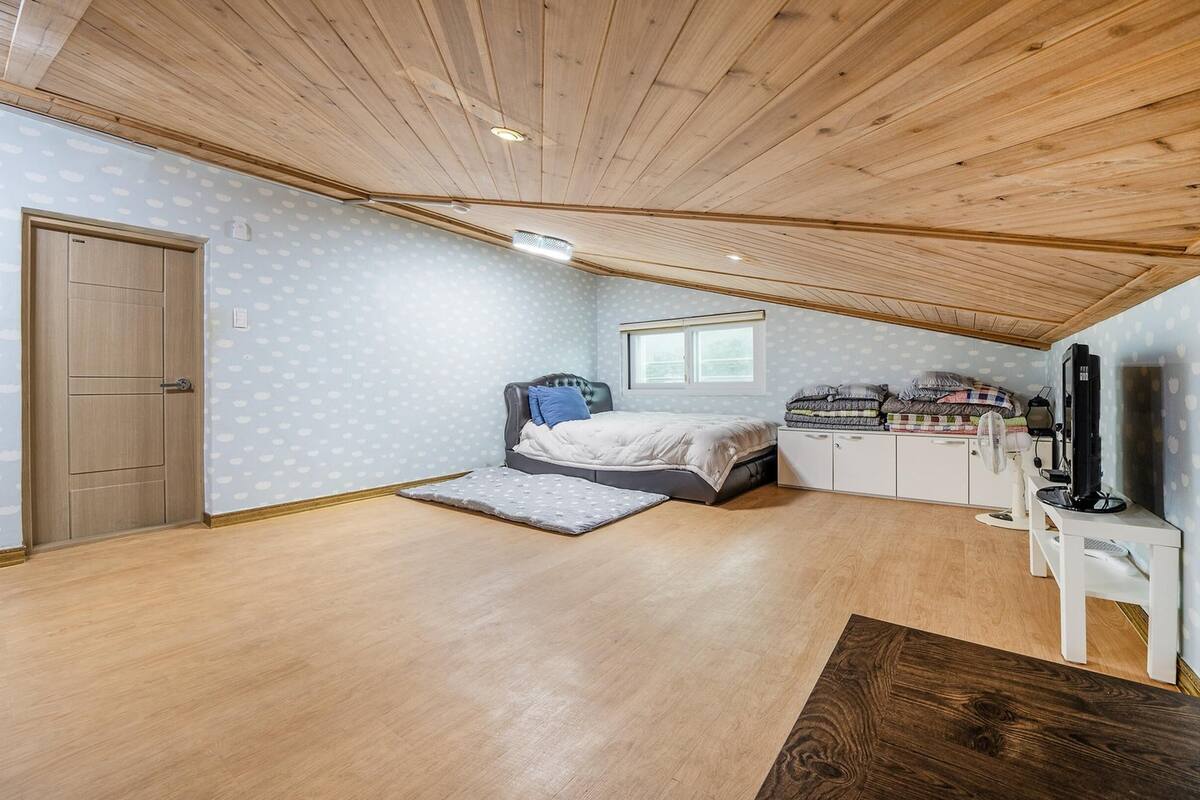 Een slaapkamer met een houten plafond, een bed met een grijze hoofdbord, een nachtkastje met een lamp, een televisie en een kledingkast.