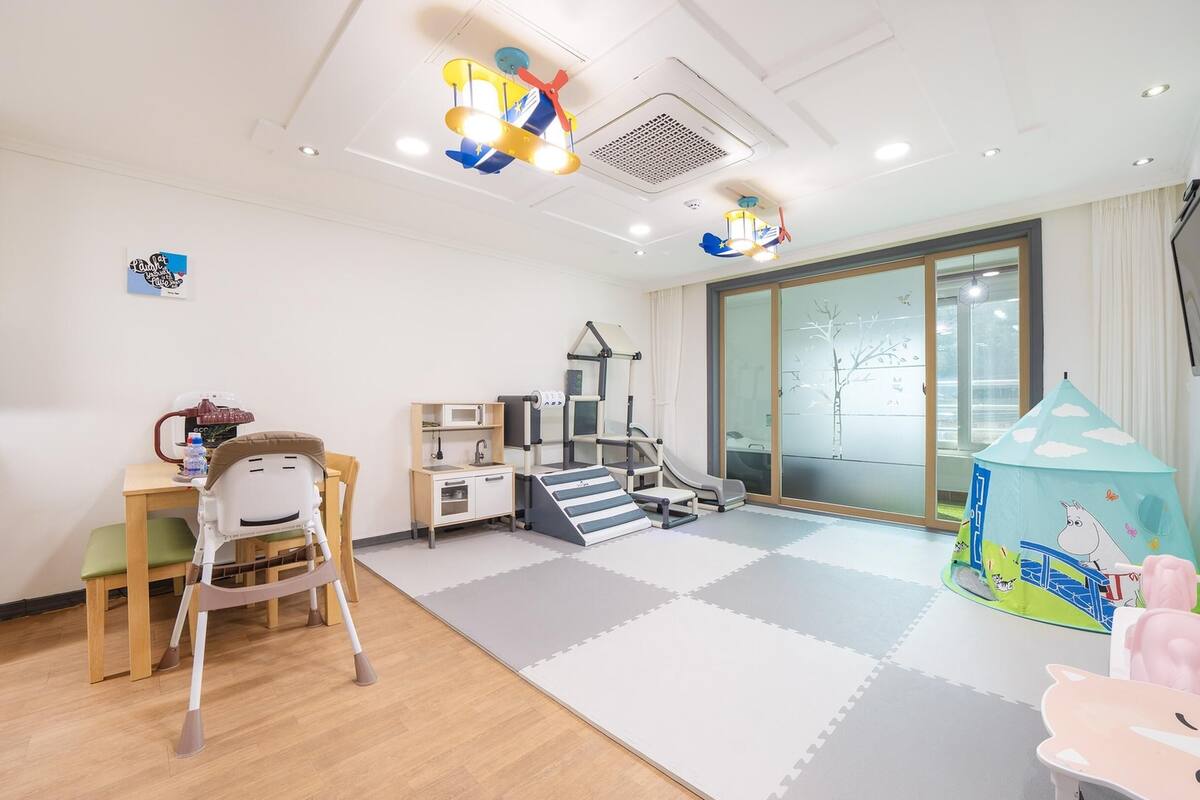 Een kindvriendelijke kamer met een speelhoekje, een klein kitchenette en een aan de muur bevestigde televisie.