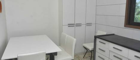 Una cocina pequeña y blanca con una mesa, sillas, un armario con cajones y un ventanal con vistas a zonas verdes.
