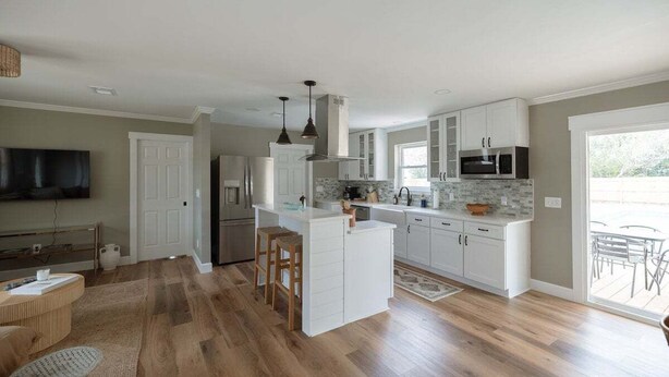 A modern kitchen with white cabinetry, a central island, and a dining area with a TV and seating. - New! Coastal Ranch with Pool and Sauna! (Wilmington)