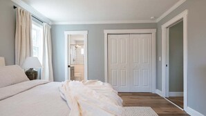 A bedroom with a large bed, a nightstand, a lamp, and a door leading to a bathroom. - New! Coastal Ranch with Pool and Sauna! (Wilmington)