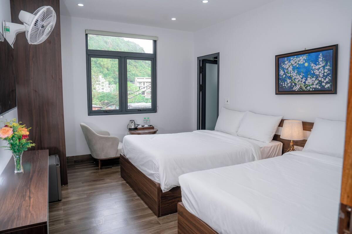 Une chambre d’hôtel avec deux lits, un bureau avec un ventilateur, une chaise, une fenêtre donnant sur un espace verdoyant et un tableau accroché au mur.