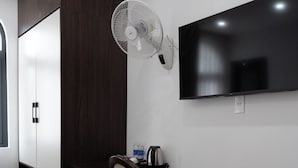 Une chambre d’hôtel avec un bureau en bois foncé, une chaise, un téléviseur fixé au mur et un ventilateur sur pied.