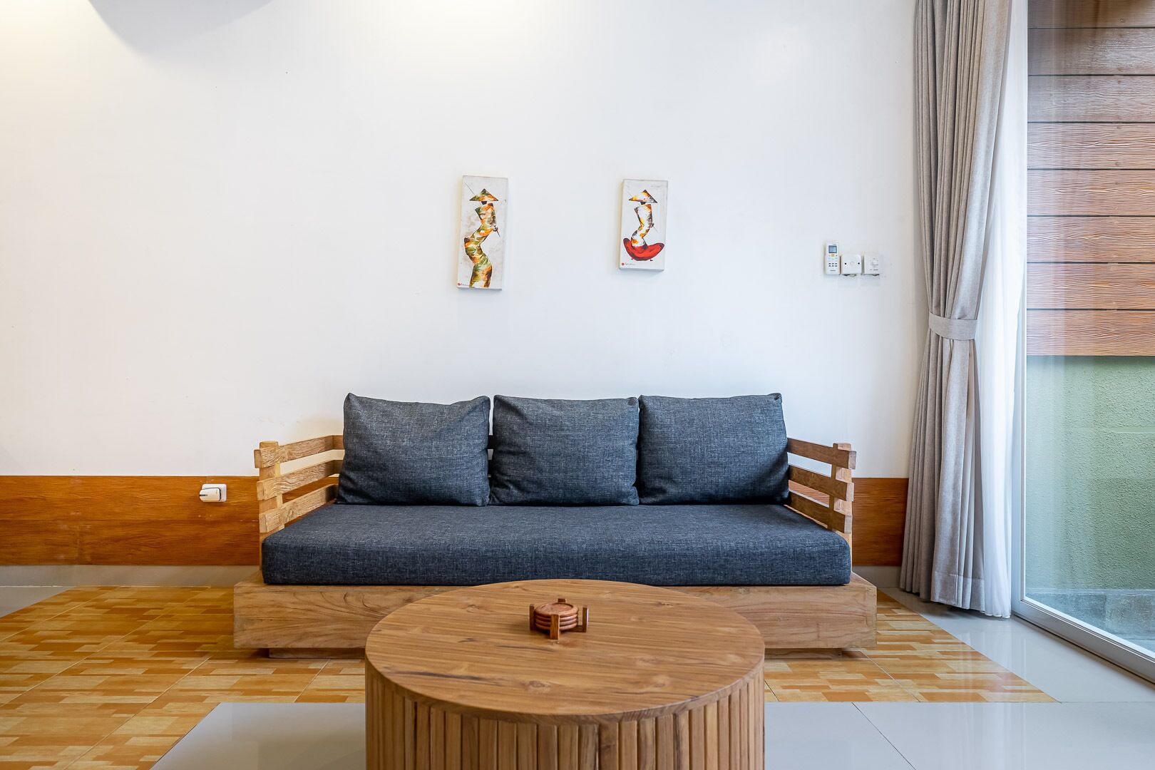 A modern living room with a grey sofa, two wall-mounted artworks, a wooden coffee table, and a large window with curtains.