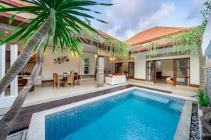 Un área de piscina exterior moderna con mobiliario de comedor, un sofá blanco y una escalera que conduce a un nivel superior cubierto.