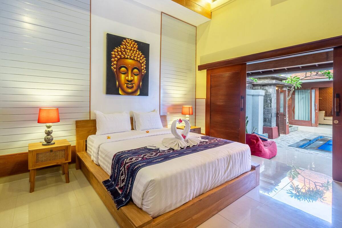 Habitación de hotel con cama de madera, dos lámparas de noche, un cuadro grande de Buda y una decoración con forma de cisne en la cama.