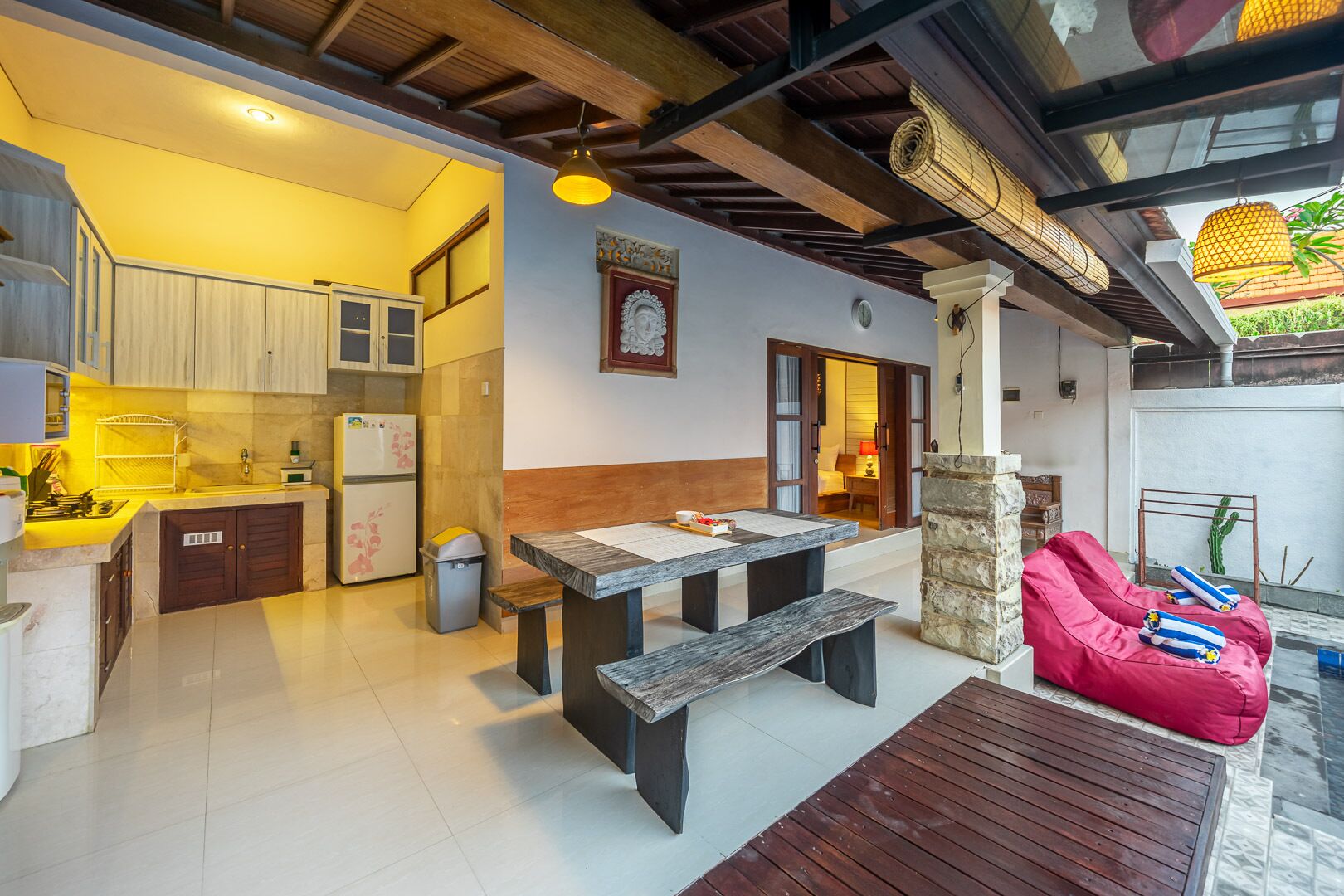 Uma cozinha compacta com eletrodomésticos modernos, um espaço de jantar com mesa e banco de madeira, e uma área de estar aconchegante com um pufe vermelho.