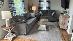 Un salon comprenant un canapé gris, un fauteuil gris plus petit, une table basse en bois, une lampe blanche, un meuble en bois et un téléviseur fixé au mur.