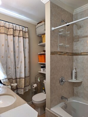 Ein Badezimmer mit Badewanne, Toilette und Waschbecken. Regale mit Toilettenartikeln und ein Duschbereich mit Handtuchhalter.