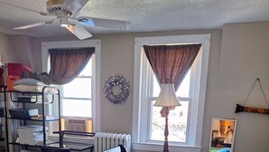 A bedroom with a bed, a desk, a radiator, a ceiling fan, and a window with curtains.