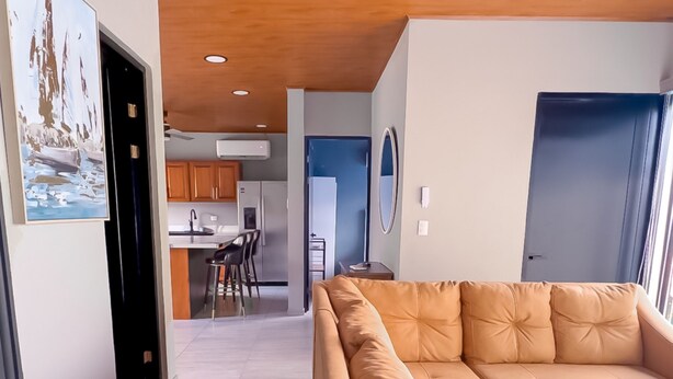 A modern living room with a beige sofa, wooden ceiling, and a kitchen in the background. - Modern Uvita Beachhouse by Whale's Tale  (Bahía Ballena)