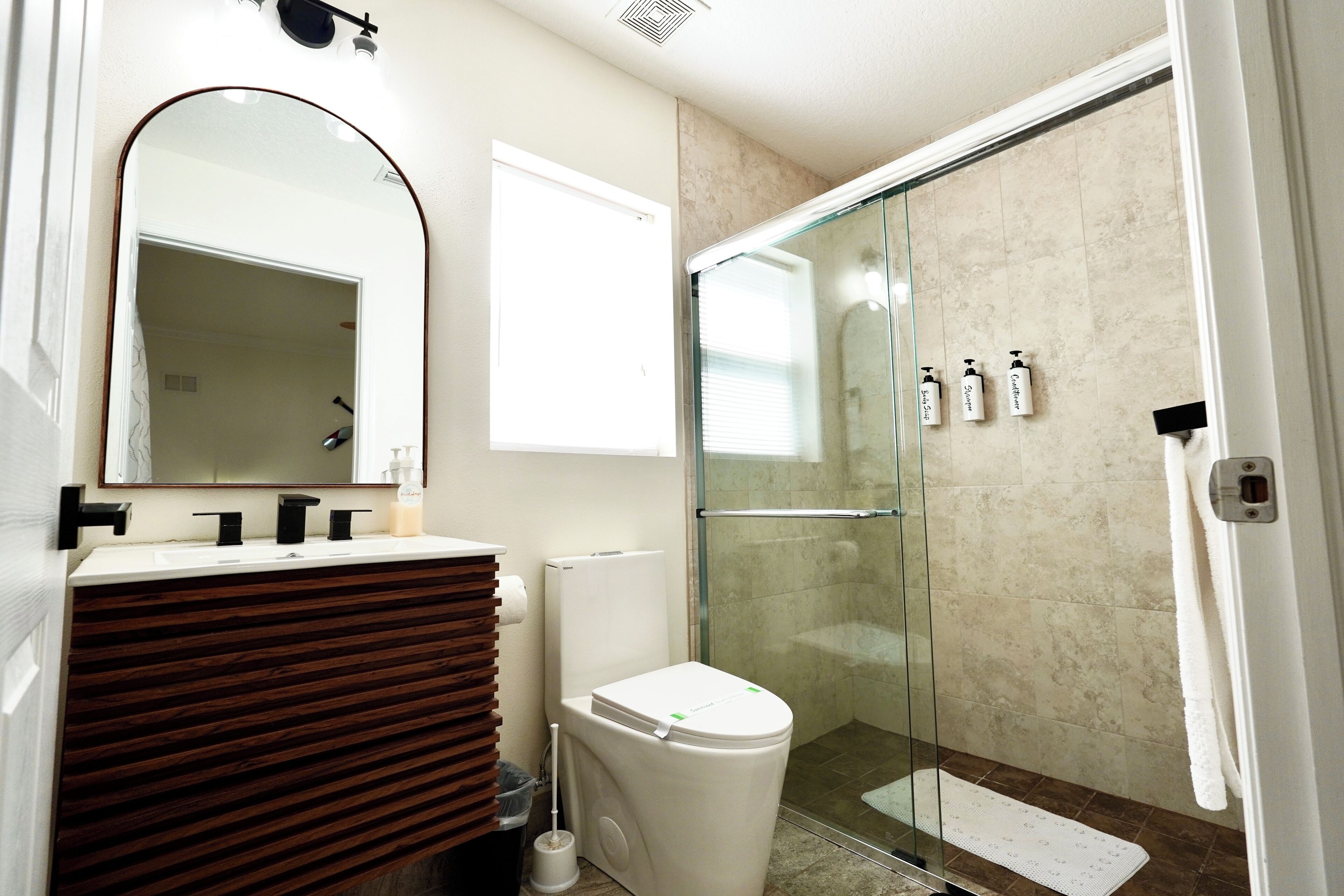 A modern bathroom with a wooden vanity, a toilet, a glass shower enclosure, and a mirror.
