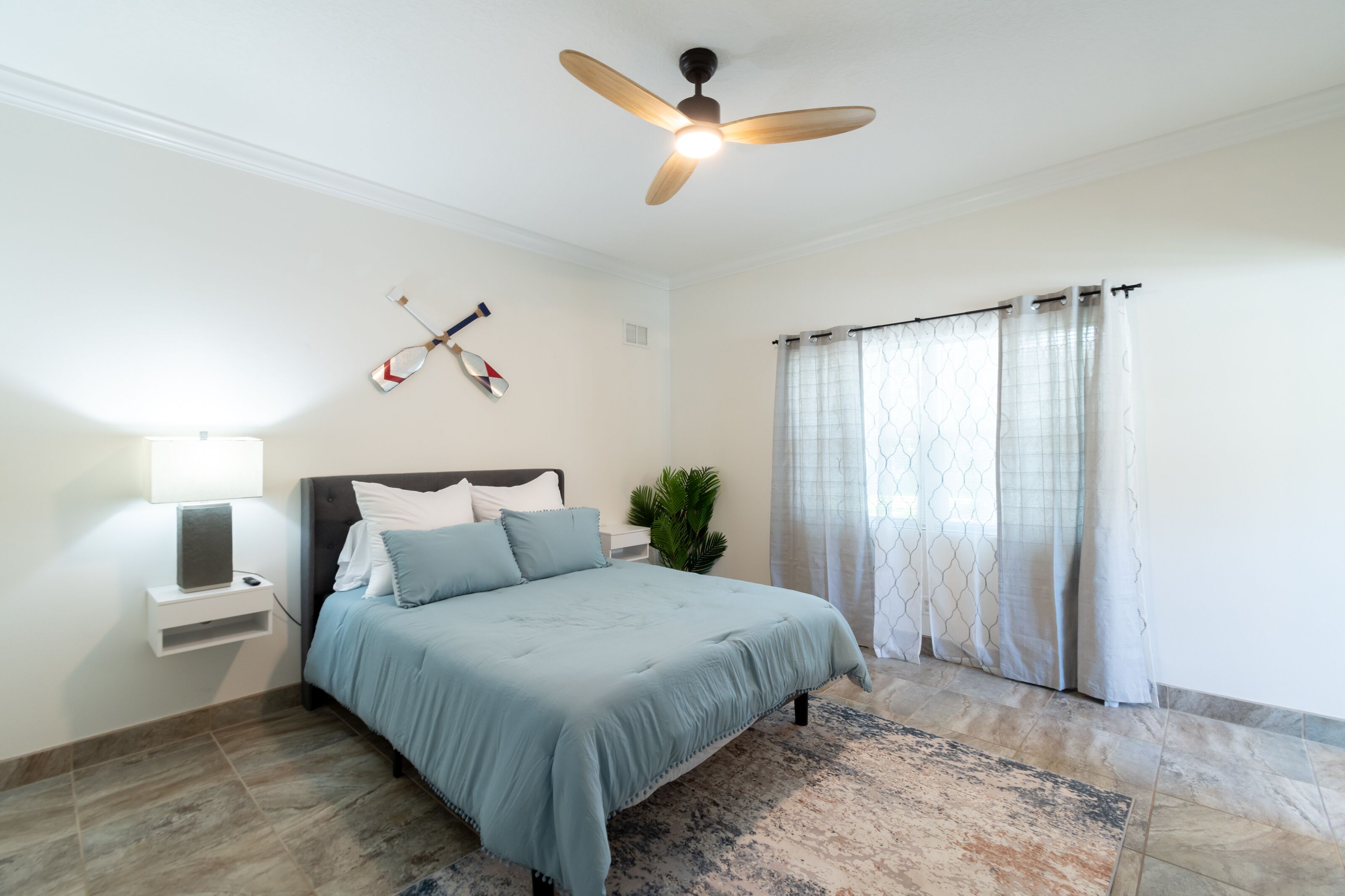 A bedroom with a bed, a ceiling fan, a nightstand, a lamp, a potted plant, and a window with curtains.