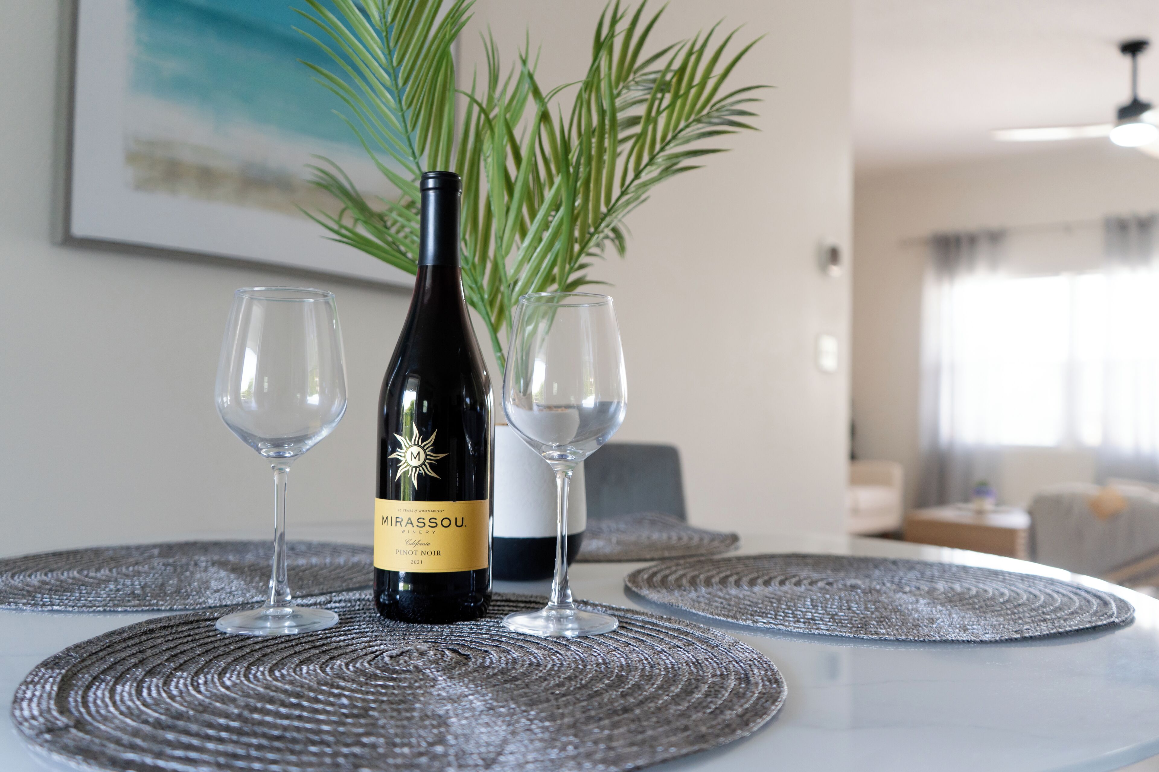 A bottle of Mirassou wine and two glasses on a table with a woven placemat, a potted plant, and a framed picture in a room with a modern design.