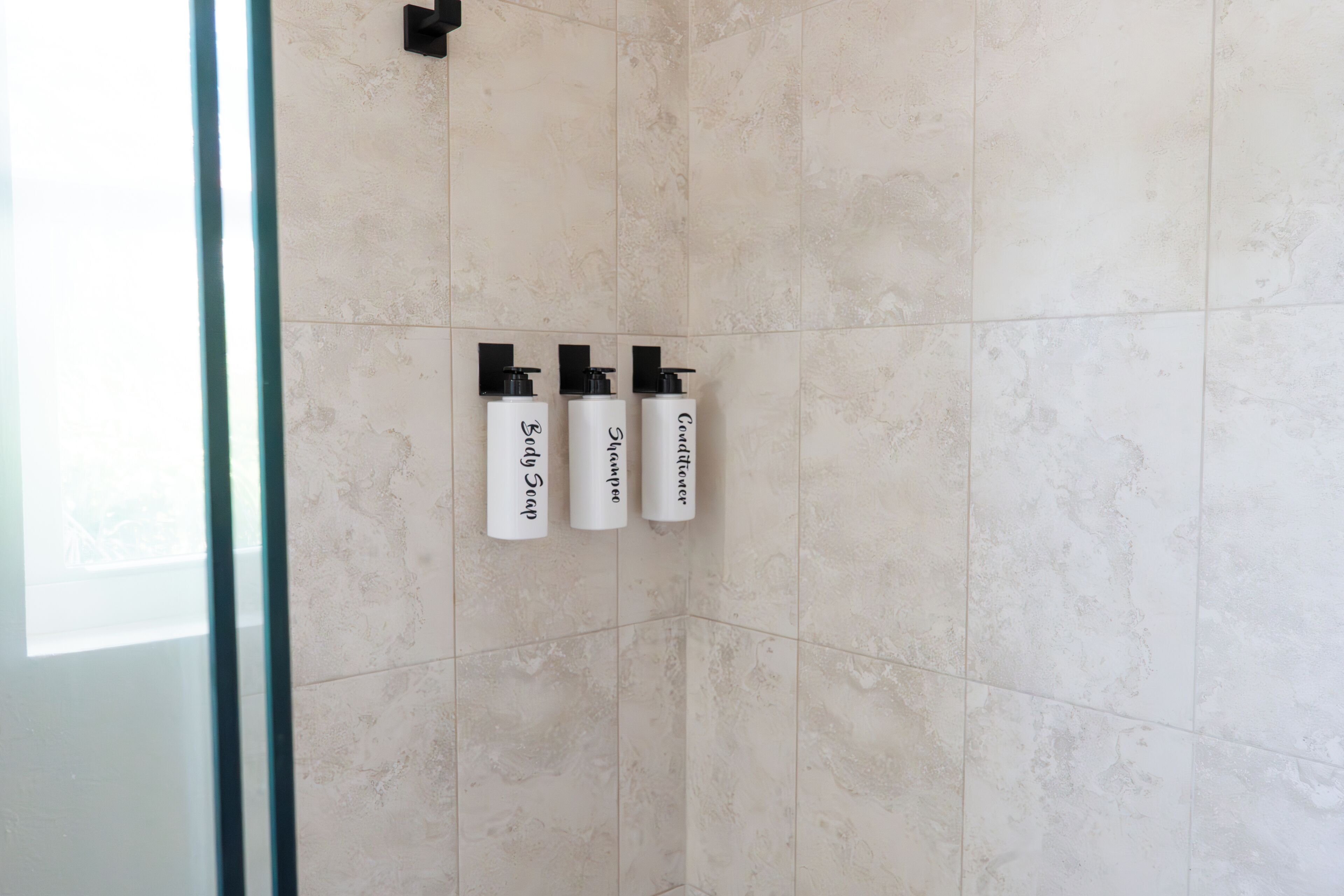 A bathroom with three white bottles labeled "Body Soap," "Shampoo," and "Conditioner" on a tiled wall.